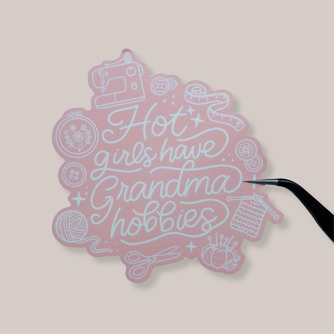 Pink sticker with text 'Hot girls have Grandma hobbies' on a beige background